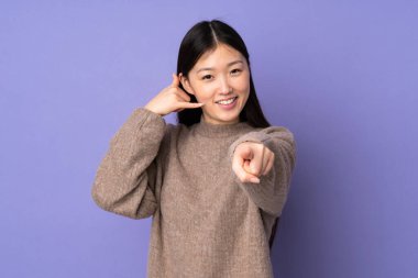 Mor arka planda izole edilmiş genç Asyalı kadın telefon hareketi yapıyor ve ön tarafı işaret ediyor.