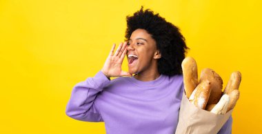 Genç Afrikalı Amerikalı bir kadın sarı arka planda izole edilmiş bir şey alıyor ve ağzı sonuna kadar açık bağırıyor.