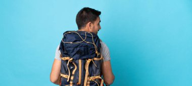 Sırt çantası ve yürüyüş direkleri olan beyaz genç bir adam mavi arka planda izole edilmiş.