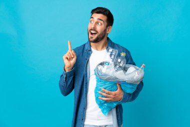 Mavi arka planda izole edilmiş plastik şişelerle dolu bir çanta tutan genç beyaz adam çözümü fark ederken parmağını kaldırmayı amaçlıyor.