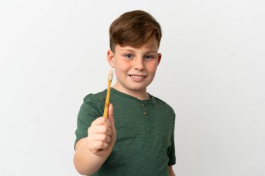 Küçük kızıl saçlı çocuk elinde diş fırçası ile beyaz arka planda mutlu bir ifadeyle izole edilmiş.