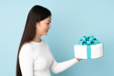 Asyalı pasta şefi elinde büyük bir pasta tutuyor mavi arka planda mutlu bir ifadeyle izole edilmiş.