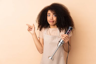 Genç Afrikalı Amerikalı kadın bej arka planda izole edilmiş el blenderi kullanıyor.