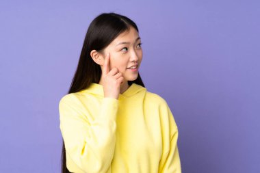 Mor arka planda izole edilmiş genç Asyalı kadın bir fikir düşünürken yukarı bakıyor.