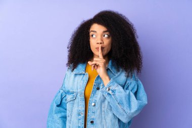 Mor arka planda izole edilmiş genç bir Afrikalı Amerikalı kadın sessiz bir jest işareti gösteriyor. Parmağını ağzına sokuyor.
