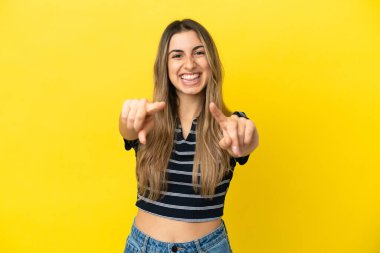 Sarı arka planda izole edilmiş genç beyaz kadın şaşırmış ve ön tarafı işaret ediyor.