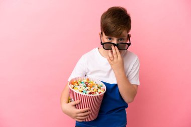 Pembe arka planda izole edilmiş küçük kızıl saçlı çocuk 3D gözlüklü ve elinde koca bir kova patlamış mısırla şaşırdı.