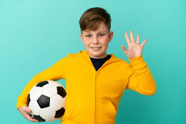 Mavi arka planda izole bir şekilde futbol oynayan kızıl saçlı çocuk mutlu bir ifadeyle selamlıyor.