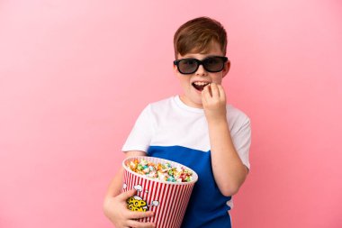 Küçük kızıl saçlı çocuk pembe arka planda 3 boyutlu gözlüklü ve elinde patlamış mısır dolu bir kova tutuyor.