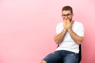 İzole edilmiş pembe arka planda bir sandalyede oturan genç adam mutlu ve gülümseyen elleriyle ağzını kapatıyor.