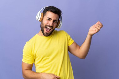 Mor arka planda sakallı genç yakışıklı adam müzik dinliyor ve gitar hareketi yapıyor.