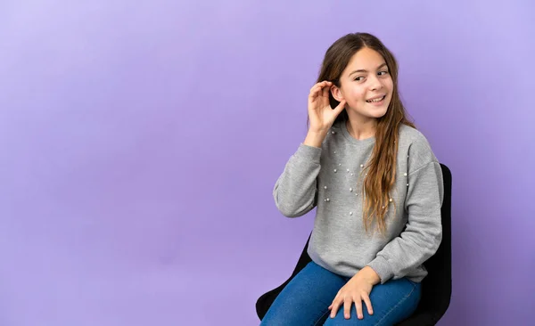 耳に手を置くことによって何かを聞いて紫色の背景に隔離された椅子に座っている小さな白人の女の子 — ストック写真