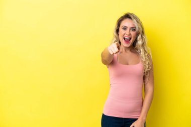 Sarı arka planda izole edilmiş genç Brezilyalı kadın şaşırmış ve ön tarafı işaret ediyor.