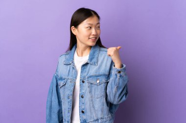 İzole edilmiş mor arka planı olan genç bir Çinli kız bir ürünü sunmak için kenarı işaret ediyor.