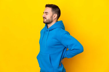 Beyaz tenli yakışıklı, sarı arka planda izole edilmiş, gayret gösterdiği için sırt ağrısı çeken genç bir adam.