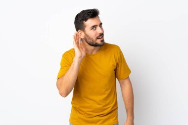 Beyaz arka planda sakallı yakışıklı genç adam elini kulağına koyarak bir şeyler dinliyor.