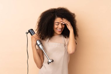 Genç Afrikalı Amerikalı kadın el blenderi kullanıyor. Bej arka planda izole edilmiş, gülerek.