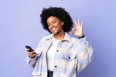 Mor arka planda izole edilmiş cep telefonu kullanan genç Afrikalı Amerikalı kadın parmaklarıyla tamam işareti gösteriyor.