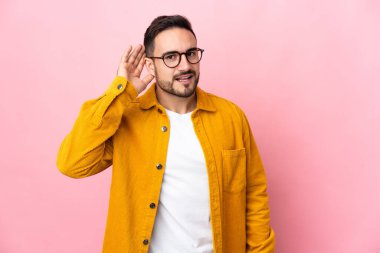 Beyaz tenli yakışıklı adam pembe arka planda izole edilmiş bir şekilde elini kulağına koyarak bir şeyler dinliyor.