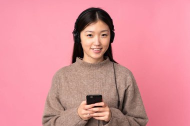 Pembe arka planda izole edilmiş genç Asyalı kadın cep telefonuyla müzik dinliyor ve ön tarafa bakıyor.