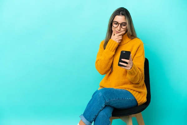 Kafkasyalı genç bir kadın mavi arka planda izole edilmiş bir sandalyede oturuyor düşünüyor ve bir mesaj gönderiyor.