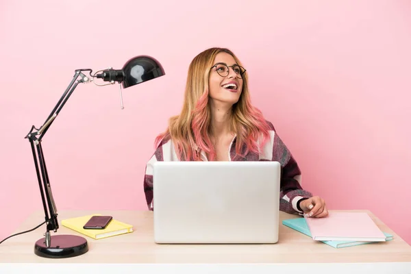 Young student woman in a workplace with a laptop over pink background laughing