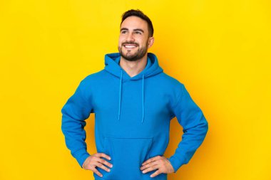 Sarı arka planda izole edilmiş, kolları kalçasında ve gülümseyen genç beyaz bir adam.