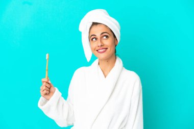 Bornozlu genç bir kadın mavi arka planda dişlerini fırçalıyor gülümserken yukarı bakıyor.