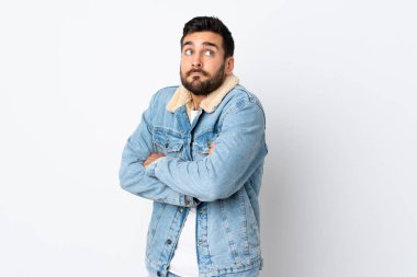 Beyaz arka planda sakallı yakışıklı genç adam, omuzlarını kaldırırken şüpheyle hareket ediyor.