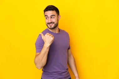 Sarı arka planda izole edilmiş beyaz, yakışıklı genç bir adam bir ürün sunmak için kenarı işaret ediyor.