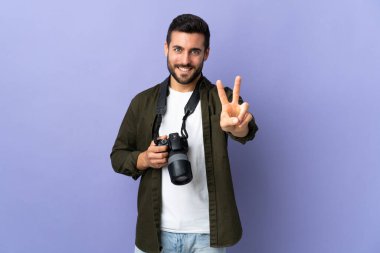 İzole edilmiş mor arka planda gülümseyen ve zafer işareti gösteren fotoğrafçı.