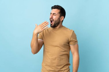 Sakallı genç, yakışıklı bir adam izole edilmiş arka planda esniyor ve ağzı açık bir şekilde elleriyle ağzını kapatıyor.