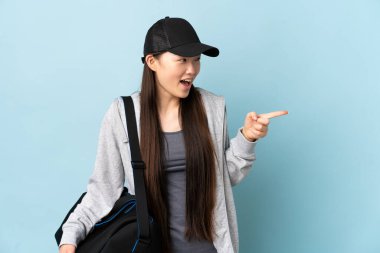 İzole edilmiş mavi arka planda spor çantasıyla genç Çinli kadın parmağını yana uzattı ve bir ürün sundu.