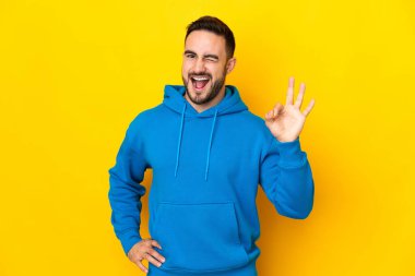 Sarı arka planda izole edilmiş beyaz, yakışıklı genç bir adam parmaklarıyla tamam işareti gösteriyor.