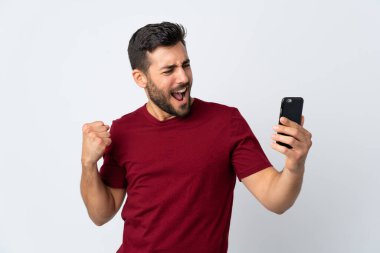 Galibiyeti kutlamak için beyaz arka planda izole edilmiş cep telefonu kullanan sakallı yakışıklı bir genç.