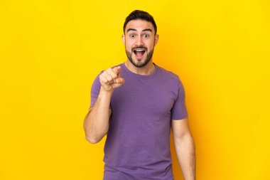 Sarı arka planda izole edilmiş beyaz, yakışıklı genç adam şaşırmış ve ön tarafı işaret ediyor.