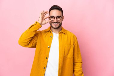 Pembe arka planda izole edilmiş beyaz, yakışıklı genç bir adam parmaklarıyla tamam işareti gösteriyor.