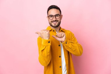 Pembe arka planda izole edilmiş beyaz, yakışıklı genç bir adam bir ürün sunmak için kenarı işaret ediyor.