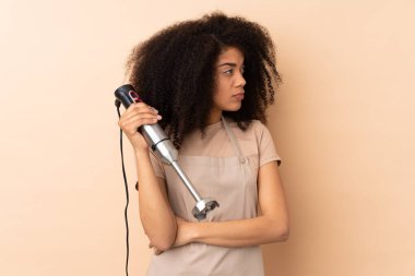 Genç Afrikalı Amerikalı kadın el blenderi kullanıyor. Bej arka planda izole edilmiş.