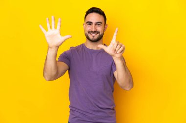 Sarı arka planda izole edilmiş beyaz, yakışıklı genç adam parmaklarıyla yediyi sayıyor.