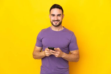 Sarı arka planda izole genç beyaz adam cep telefonuyla bir mesaj gönderiyor.