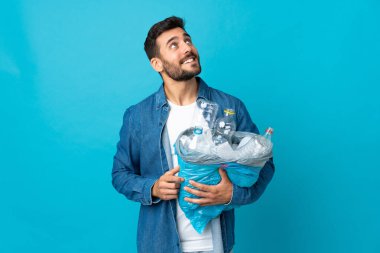 Mavi arka planda izole edilmiş plastik şişelerle dolu bir çanta taşıyan genç beyaz adam gülümserken yukarı bakıyor.
