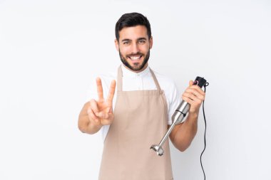 Beyaz arka planda izole edilmiş el blenderi kullanan adam gülümsüyor ve zafer işareti gösteriyor.