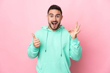Pembe arka planda izole edilmiş beyaz, yakışıklı genç bir adam tamam işareti ve baş parmak hareketi gösteriyor.