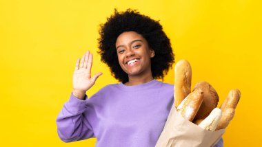 Genç Afrikalı Amerikalı bir kadın sarı arka planda izole edilmiş bir şey alıyor mutlu bir ifadeyle selamlıyor.