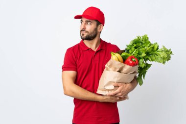 Sakallı genç adam elinde sebze dolu bir çanta beyaz arka planda izole edilmiş.