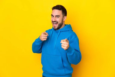 Sarı arka planda izole edilmiş beyaz, yakışıklı genç adam şaşırmış ve ön tarafı işaret ediyor.