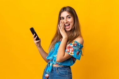 İzole edilmiş sarı arka plan üzerinde cep telefonu kullanan genç bir kadın fısıldıyor: