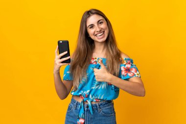 İzole edilmiş sarı arka planda cep telefonu kullanan genç bir kadın. Başparmakları havada çünkü iyi bir şey oldu.