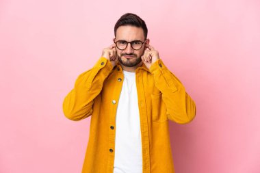 Pembe arka planda izole edilmiş genç beyaz yakışıklı adam hayal kırıklığına uğramış ve kulaklarını kapatmış.